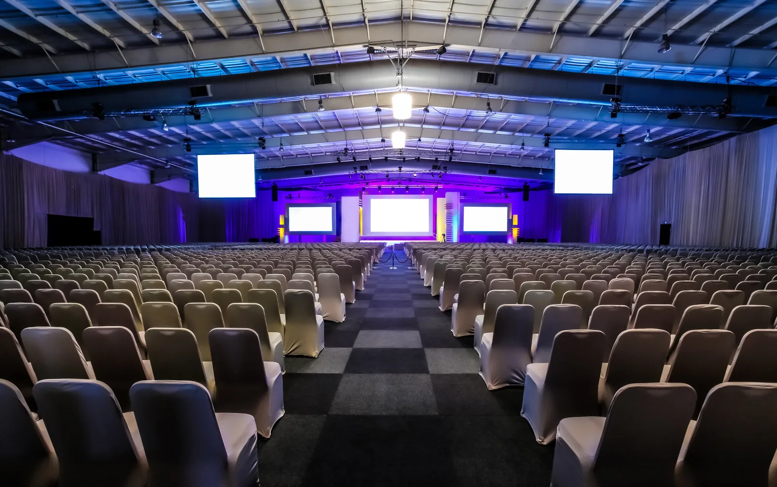 Large conference hall set for a general session — rows of dressed chairs facing a stage with three screens under blue and violet wash