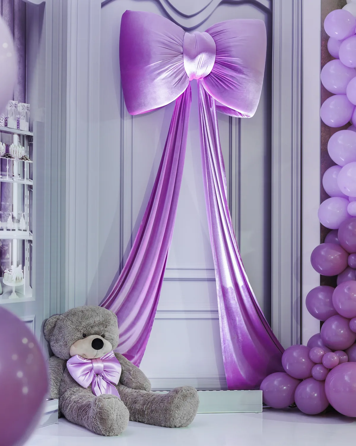 Event decor build in lilac and white — balloon columns, a draped satin bow backdrop and a dessert display, with a large teddy bear at the base