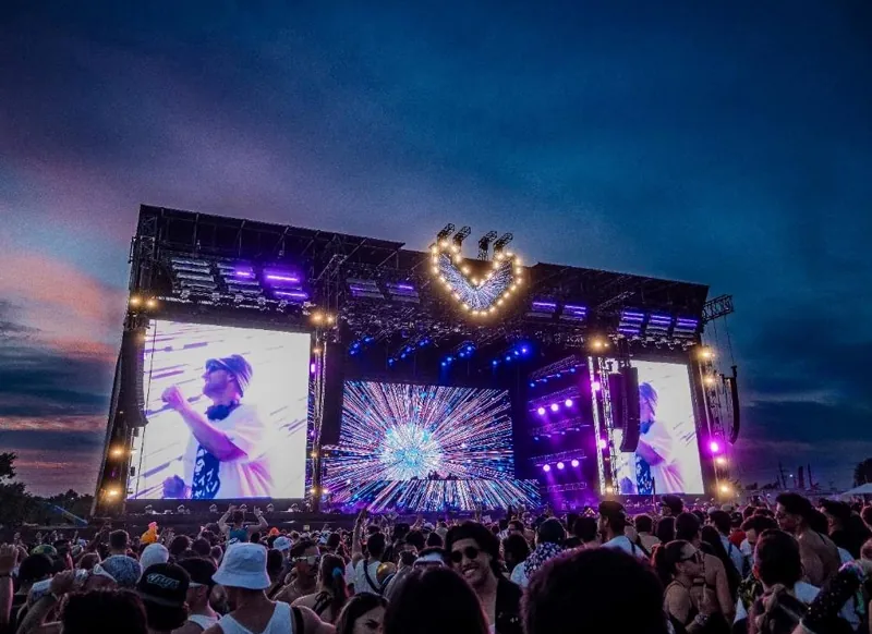 Outdoor festival stage at dusk — LED screens either side of a lit centre structure, crowd in front