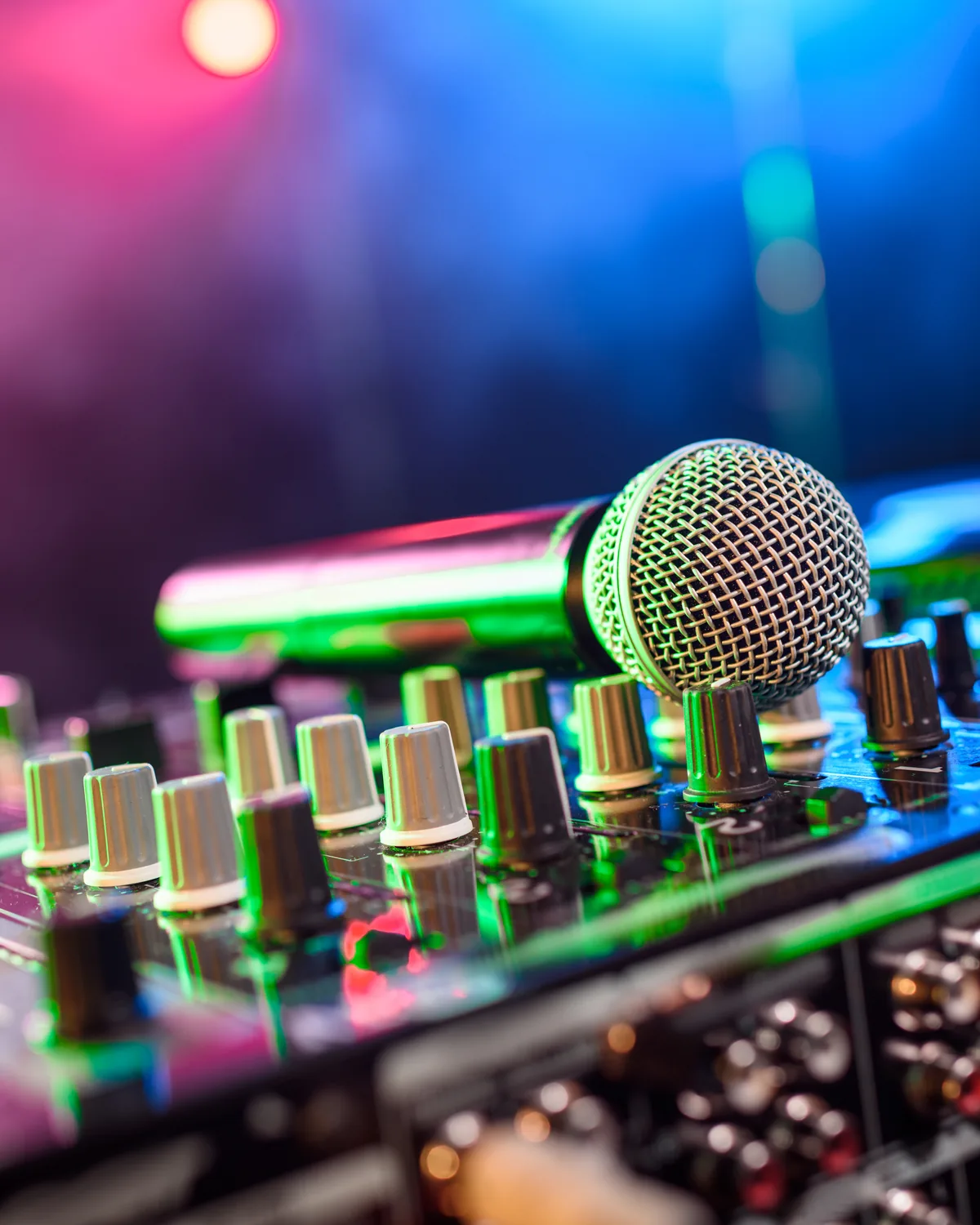 Close-up of a mixing desk with a wireless microphone resting on it, lit by pink and blue stage lighting