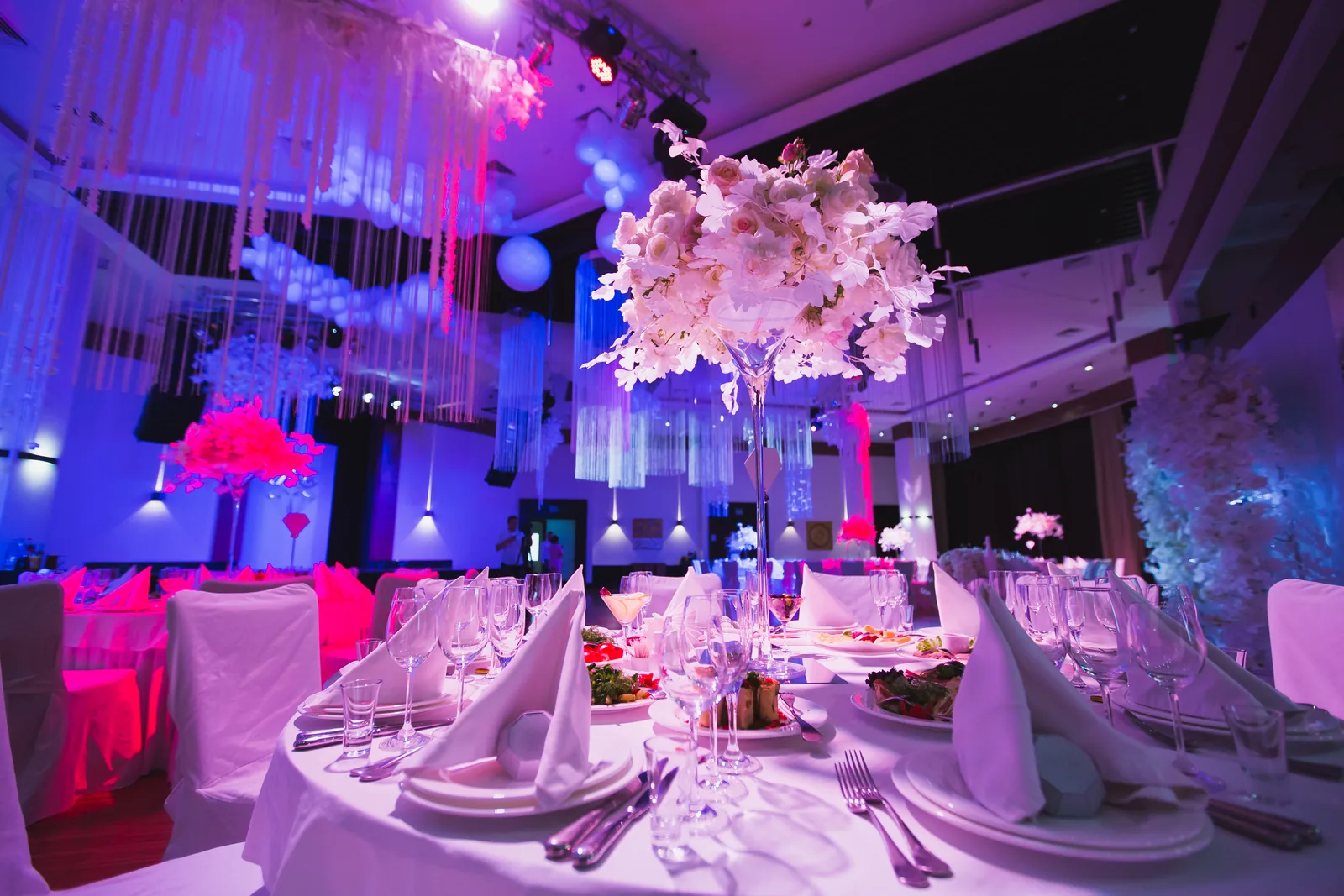 Private event ballroom with tall floral centrepieces on dressed tables, the room uplit in magenta and blue