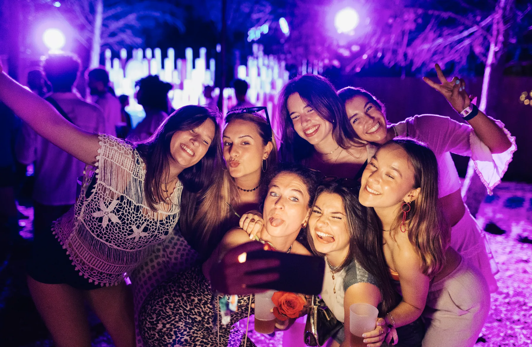 Guests taking a group selfie at a sweet sixteen party under purple and magenta event lighting, with an illuminated backdrop behind them